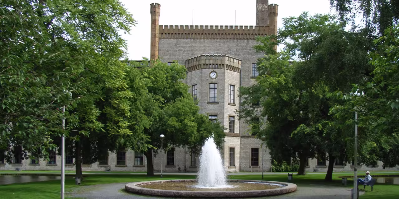 Die Ravensberger Spinnerei in der Frontalansicht. Links und rechts große Bäume. Im Fokus ein Springbrunnen direkt vor dem historischen Gebäude.