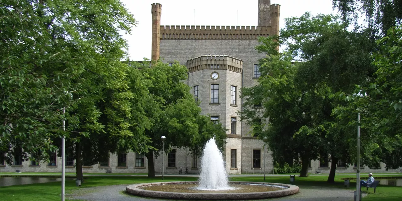 Die Ravensberger Spinnerei in der Frontalansicht. Links und rechts große Bäume. Im Fokus ein Springbrunnen direkt vor dem historischen Gebäude.