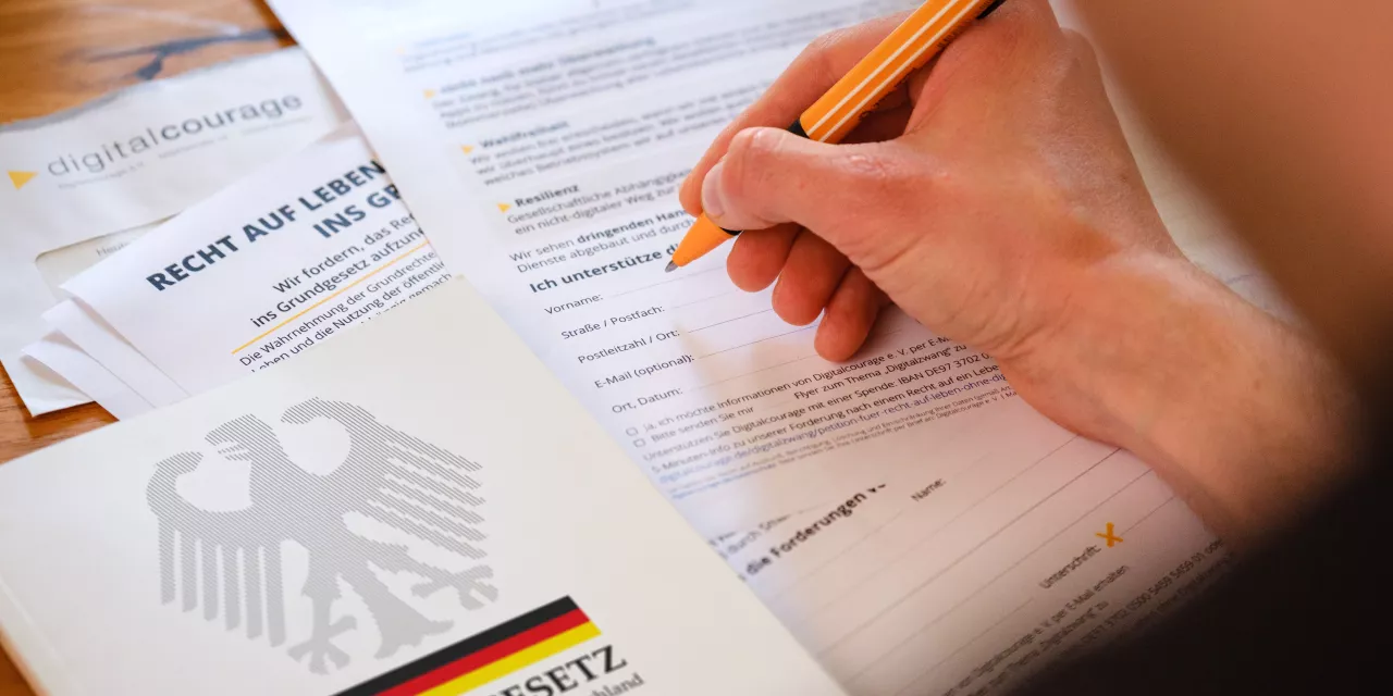 Eine Person ist dabei, die Petition zu unterzeichnen. Neben der ausgedruckten Petition liegt das Grundgesetz.