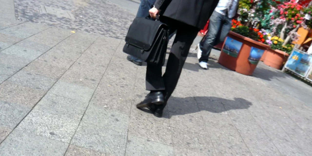 Eine Geschäftsperson im Anzug mit schwarzem Aktemkoffer auf einer Straße. Nur der Beinbereich ist zu sehen.