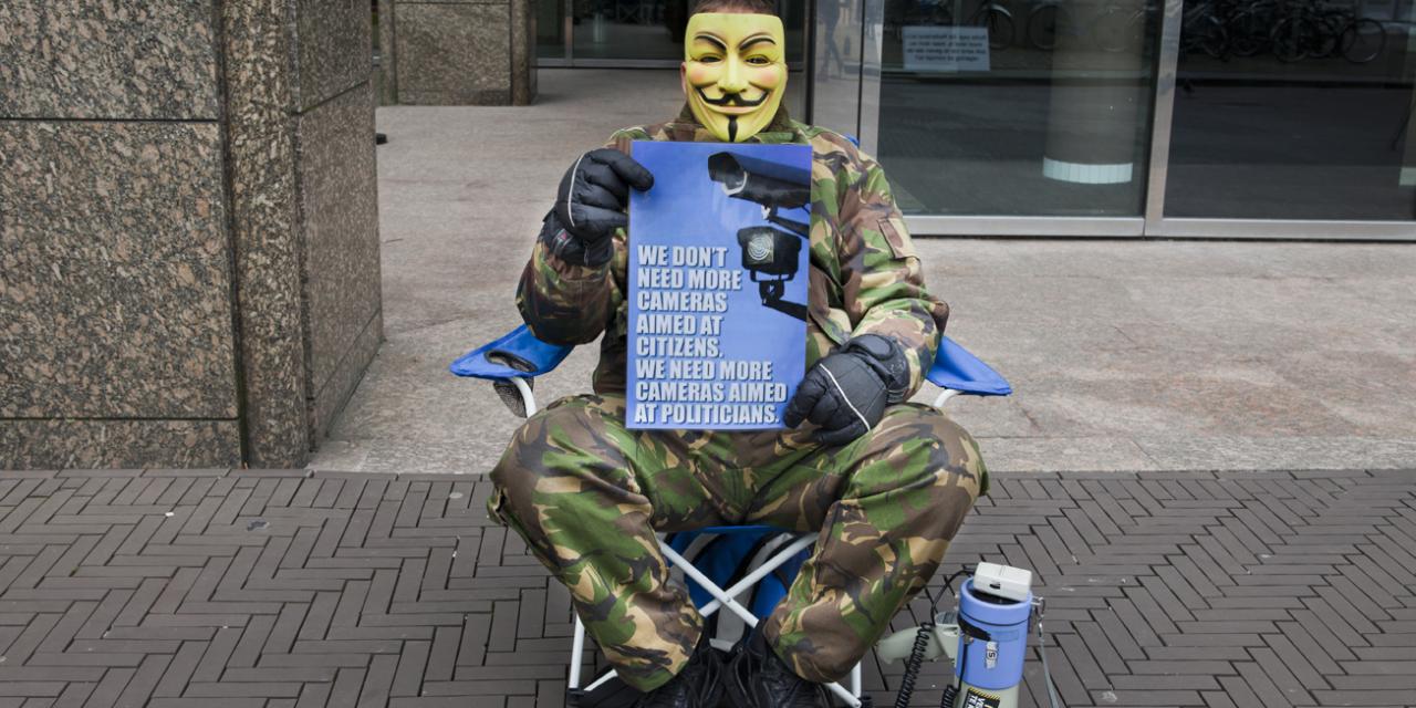 Eine Person in Trananzug und einer Guy-Fawkes-Maske sitzt auf einem Stuhl. In der Hand ein Demo-Plakat.