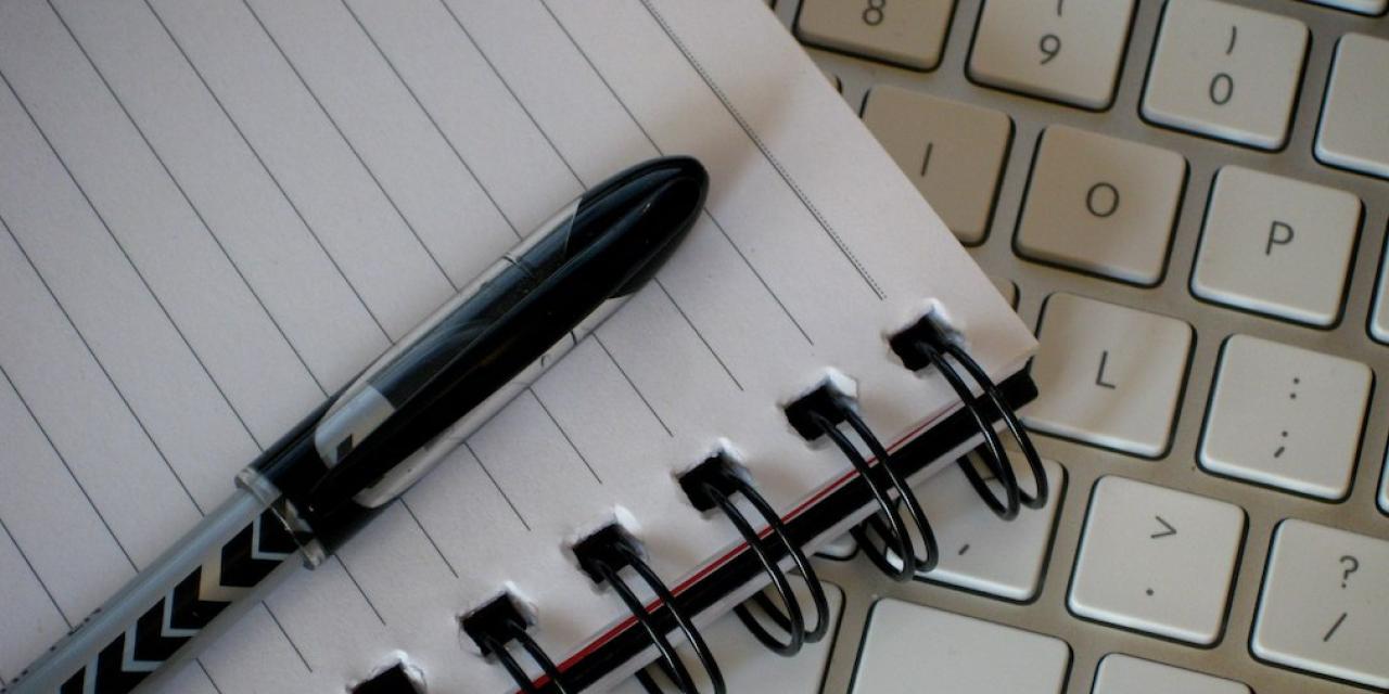 Ein Notizblock mit einem Stift. Darunter eine Tastatur.