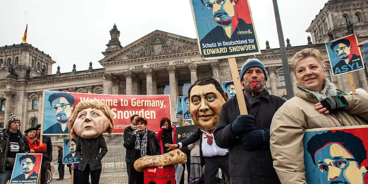 Aktivist.innen mit diversen Bannern. Im Hintergrund Pappmaché-Köpfe von Angela Merkel und Sigmar Gabriel.