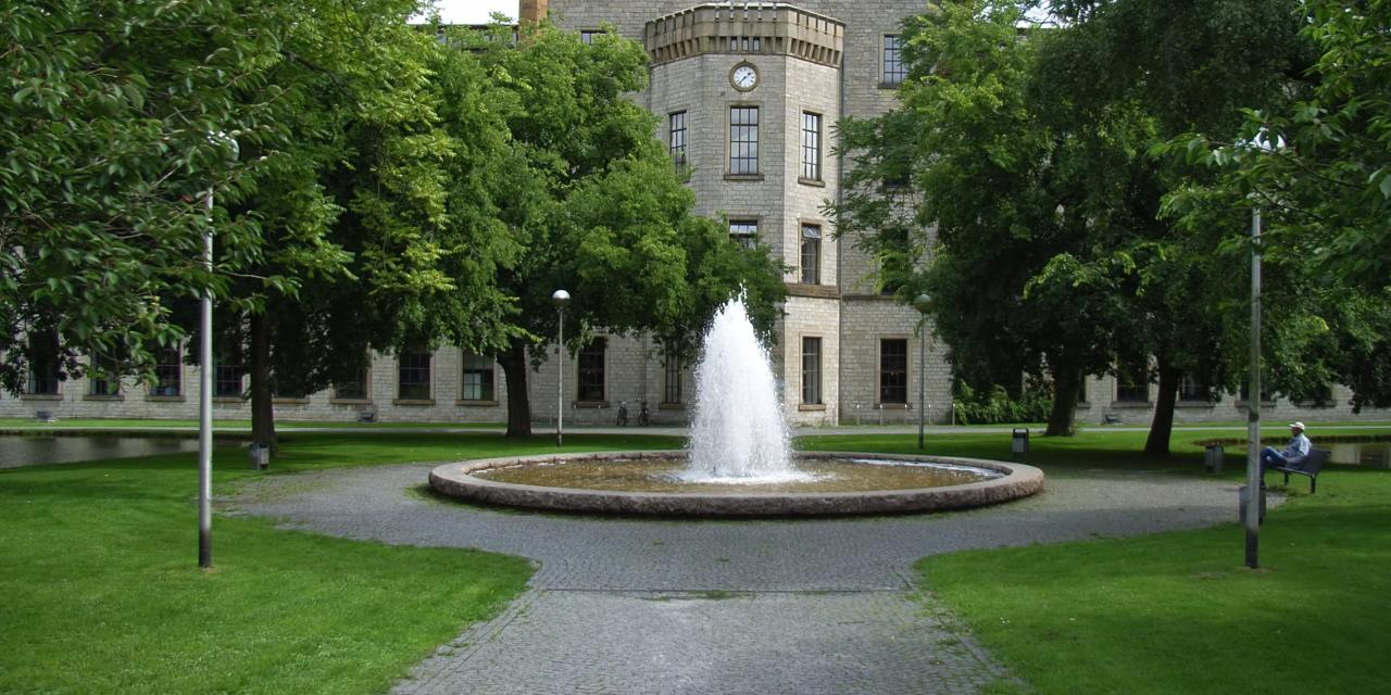 Die Ravensberger Spinnerei in der Frontalansicht. Links und rechts große Bäume. Im Fokus ein Springbrunnen direkt vor dem historischen Gebäude.