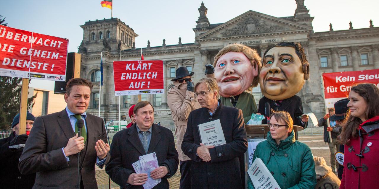 Aktivist.innen mit diversen Bannern. Im Hintergrund Pappmaché-Köpfe von Angela Merkel und Sigmar Gabriel.