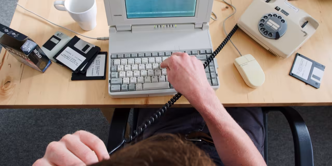 Draufsicht wie eine Person auf einem sehr alten Laptop tippt. Daneben Disketten und ein Wählscheibentelefon.