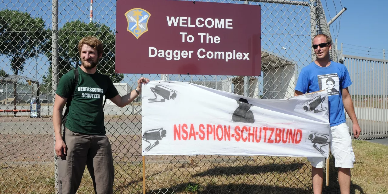 Zwei Personen halten ein Banner mit der Aufschrift: „NSA-Spion-Schutzbund“.