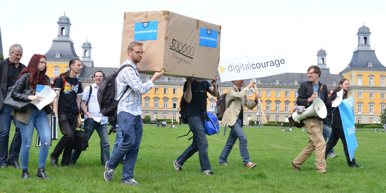 Mehrere Personen tragen ein riesiges Paket über eine Wiese. Auf der Box steht 500.000 Eingaben.