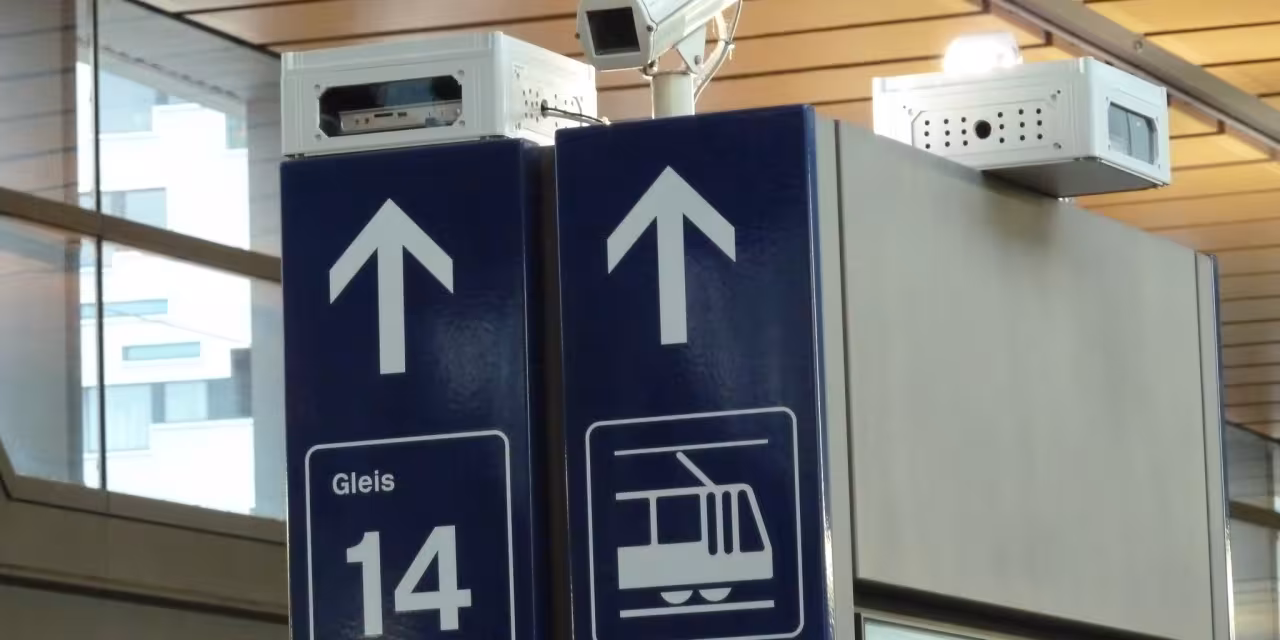 Detailaufnahmer von Hinweisschildern am Bahnhof. Darüber eine Überwachungskamera.