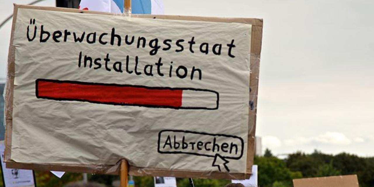 Demoschild mit einem Ladebalken und dem Text „Überwachungsstaat Installation“. Darunter ein Mauszeiger, der auf „Abbrechen“ klickt.