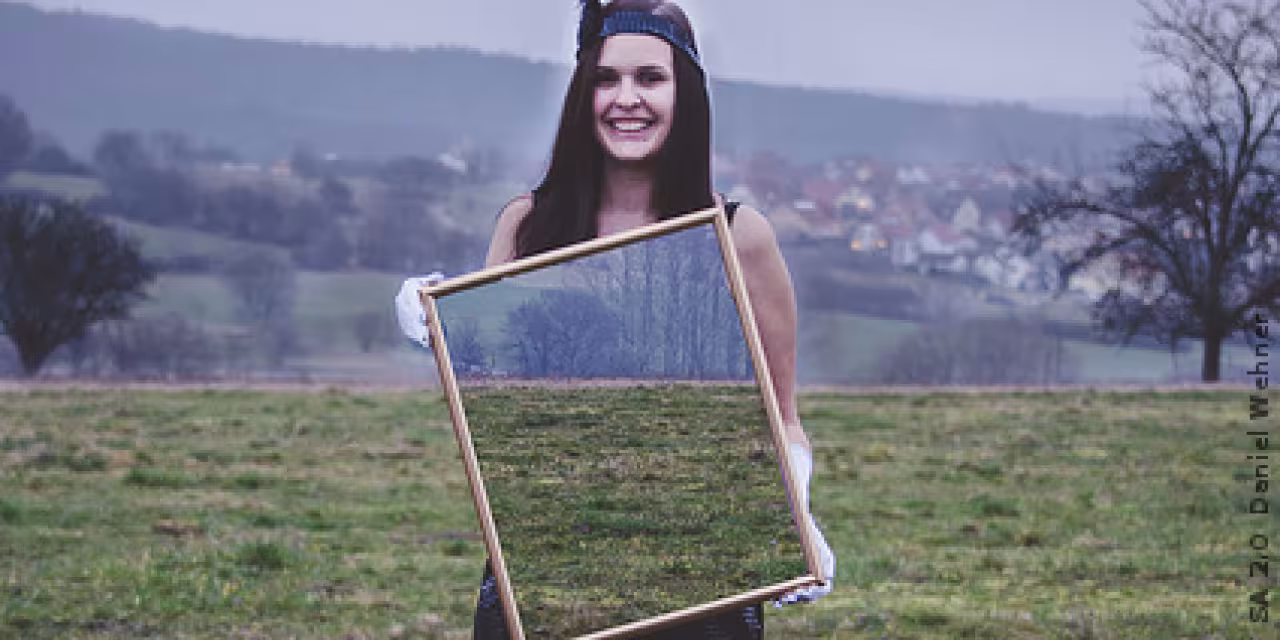 Eine Frau steht auf einem Feld und hält einen großen Spiegel vor ihrem Torso in ihren Händen. Es spiegelt nicht sich die Kamera, sondern es erscheint das Feld hinter ihr.