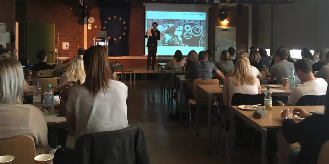 Schüler.innen an Tischen in einer Klasse. Auf einem Podium Friedemann Ebelt. Das Beamer-Bild zeigt das Logo von Digitalcourage und eine Grafik eines Kopfes bestehend aus Zahnrädern.