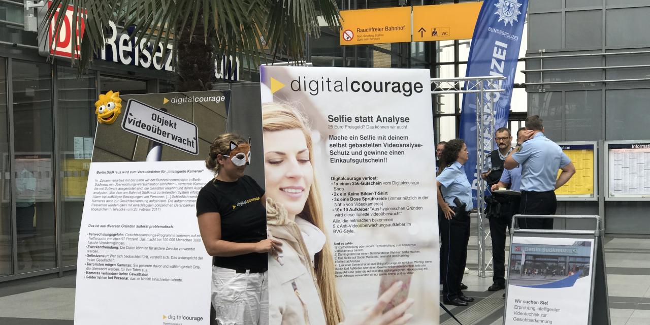 Leena Simon mit einer Maske neben einem Plakat „Selfie statt Analyse“. Im Hintergrund sind mehrere Polizist.innen. Aufnahmeort: Ein Bahnhof.