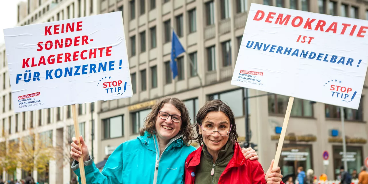 Zwei Demonstrant.innen mit Schildern, auf denen steht: „Keine Sonderklagerecht für Konzerne“ und „Demokratie ist unverhandelbar“.