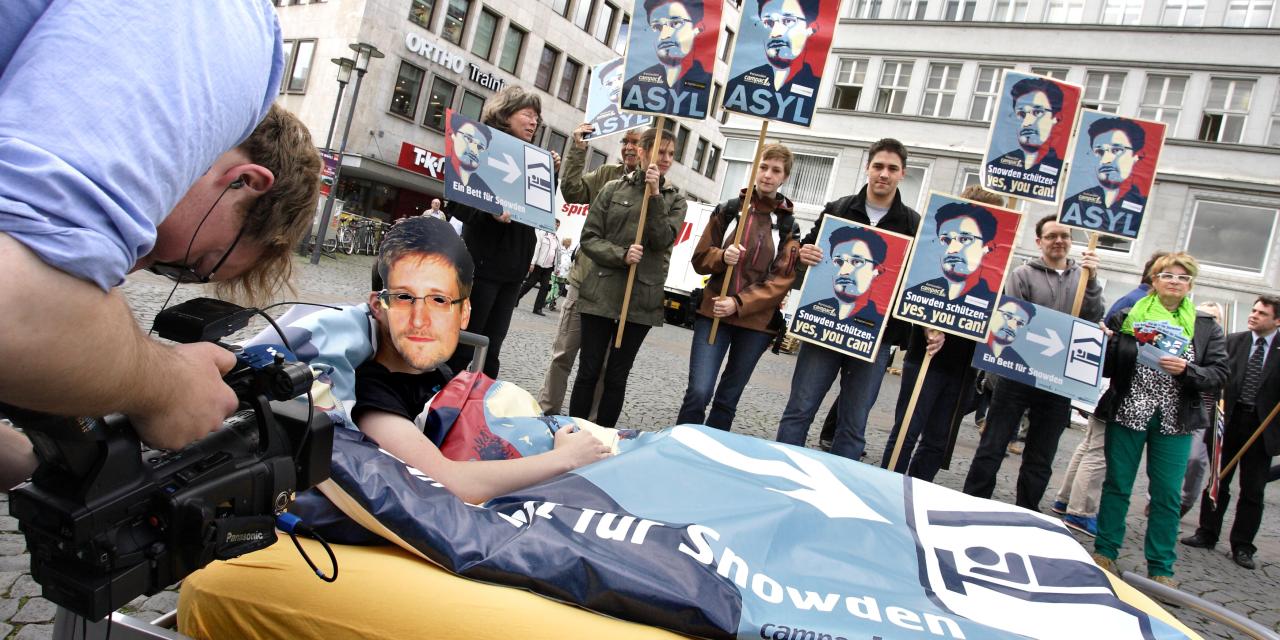 Aktion in der Innenstadt: Eine Person mit Edward-Snowden-Maske liegt in einem Bett. Im Hintergrund Menschen mit Edward-Snowden-Schildern.