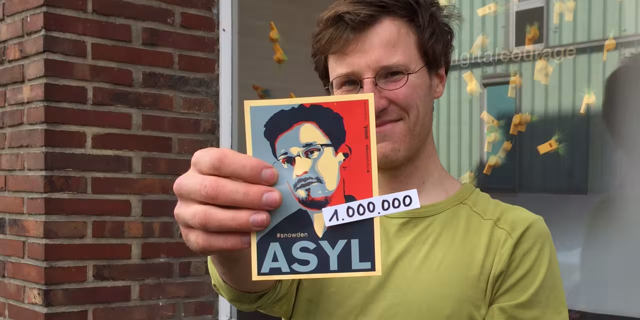 Eine Person hält einen Edward-Snowden-Asyl-Aufkleber Richtung Kamera.