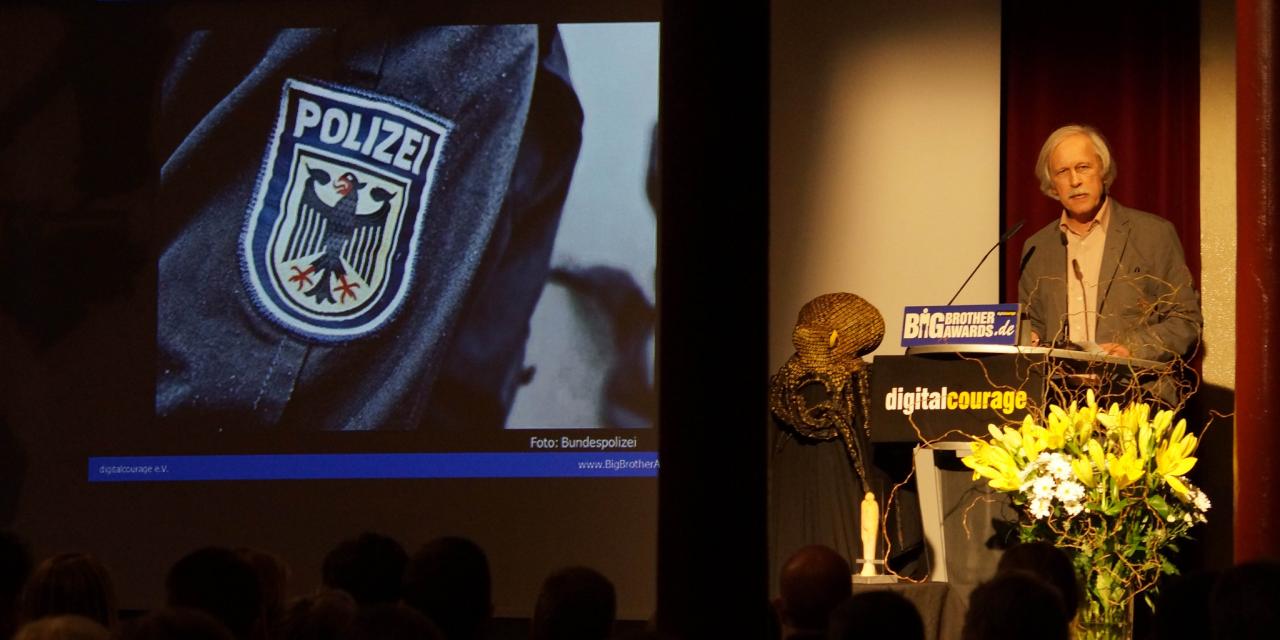 Rolf Gössner am Redepult während einer BBA-Laudatio. Beamerbild: Wappen der Polizei.