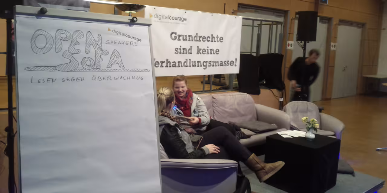 Ein Flipchart mit dem Text "Open Sofa – Lesen gegen Überwachung". Im Hintergrund zwei Personen auf einem Sofa sitzend.