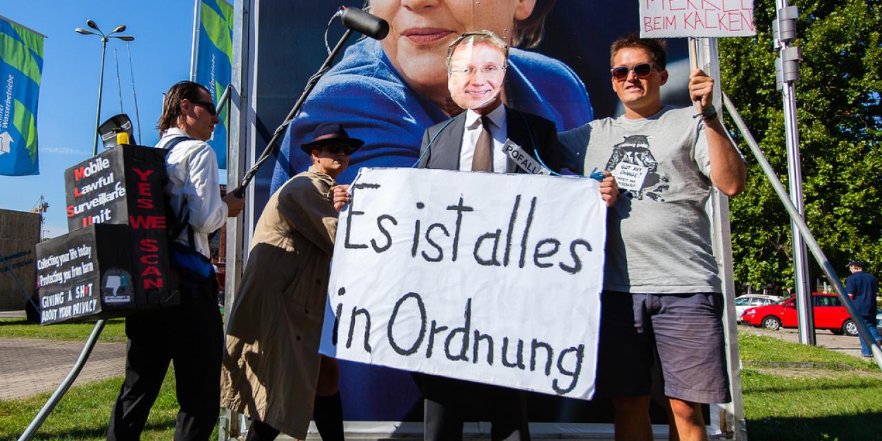 Mehrere Aktivist.innen vor einem Plakat mit Angela Merkel. Sie halten Demo-Schilder („Es ist alles in Ordnung“) und ein Mikrofon in der Hand.