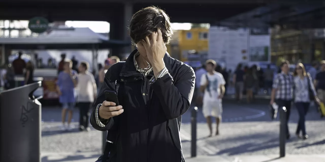 Eine Person in belebter Umgebung, der auf sein Handy schaut und sich an den Kopf fasst.