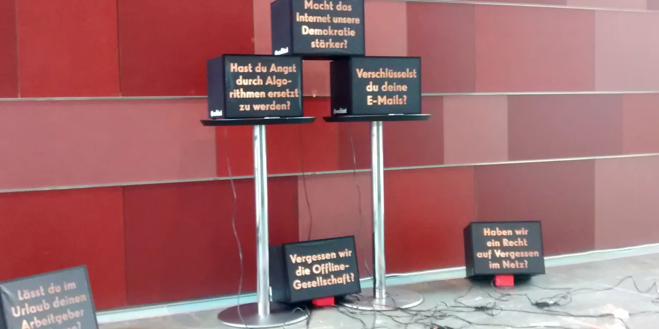 Installation: Mehrere Fernseher vor roter Wand. Darauf sind diverse Texte abgebildet.